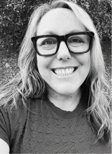 A woman with long, wavy hair and glasses smiles at the camera outdoors in this black and white photo, capturing the confidence of a digital marketing professional.