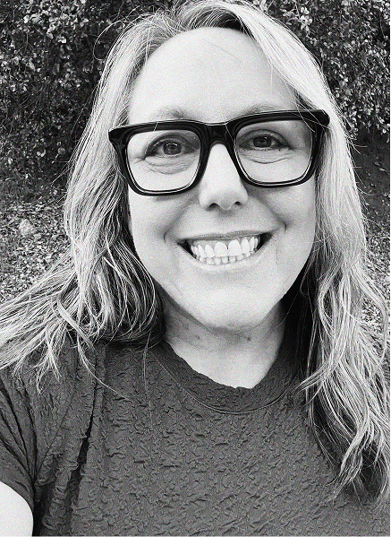 A woman with long, wavy hair and glasses smiles at the camera outdoors in this black and white photo, capturing the confidence of a digital marketing professional.