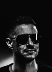 A man with short hair, wearing dark sunglasses and a slight smile, is photographed in black and white against a dark background—reflecting the modern edge of digital marketing professionals.