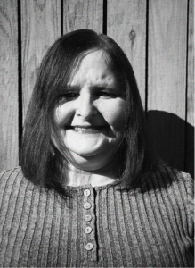 A woman with straight shoulder-length hair and a ribbed, buttoned shirt stands in front of a wooden fence, smiling in black and white—capturing the approachable face behind digital marketing and SEO success.