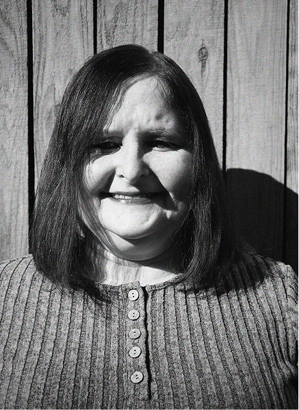 A woman with straight shoulder-length hair and a ribbed, buttoned shirt stands in front of a wooden fence, smiling in black and white—capturing the approachable face behind digital marketing and SEO success.