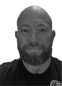 A bearded man with a bald head smiles slightly, facing forward, dressed in a dark shirt. The plain, light-colored background gives him a professional look suitable for digital marketing or CRM profiles.