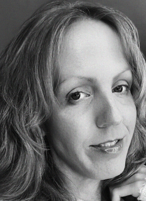 Black and white close-up photo of a woman with long wavy hair, looking at the camera with a slight smile, wearing a light-colored top.