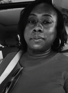 A woman with hoop earrings and a shoulder strap sits in a car, looking at the camera. Sunglasses hang from her shirt collar.