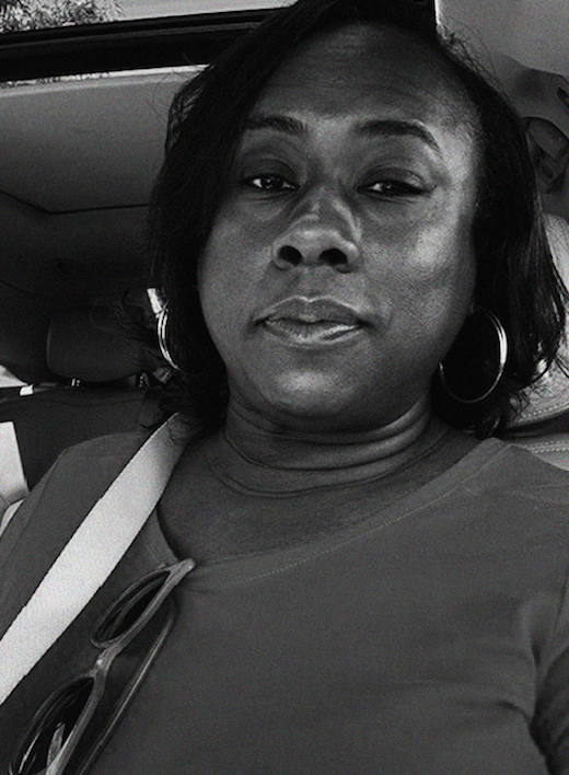 A woman with hoop earrings and a shoulder strap sits in a car, looking at the camera. Sunglasses hang from her shirt collar.